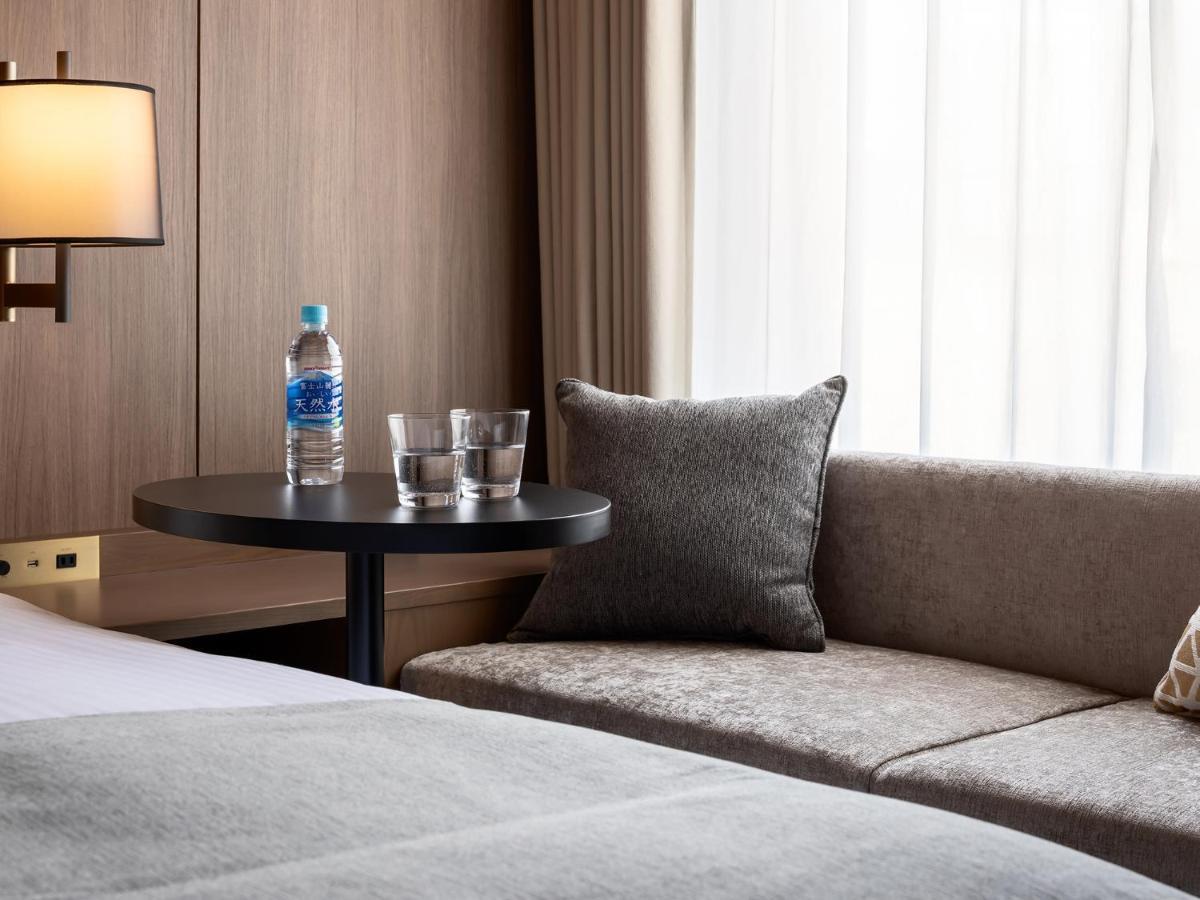 Mitsui Garden Hotel Roppongi Tokyo Premier Екстериор снимка The photo depicts a cozy hotel room setting. On the left, there is a bedside table with a lamp. Against the wall, there is a comfortable-looking couch with a decorative pillow. In front of the couch, there's a small round table that holds a bottle of