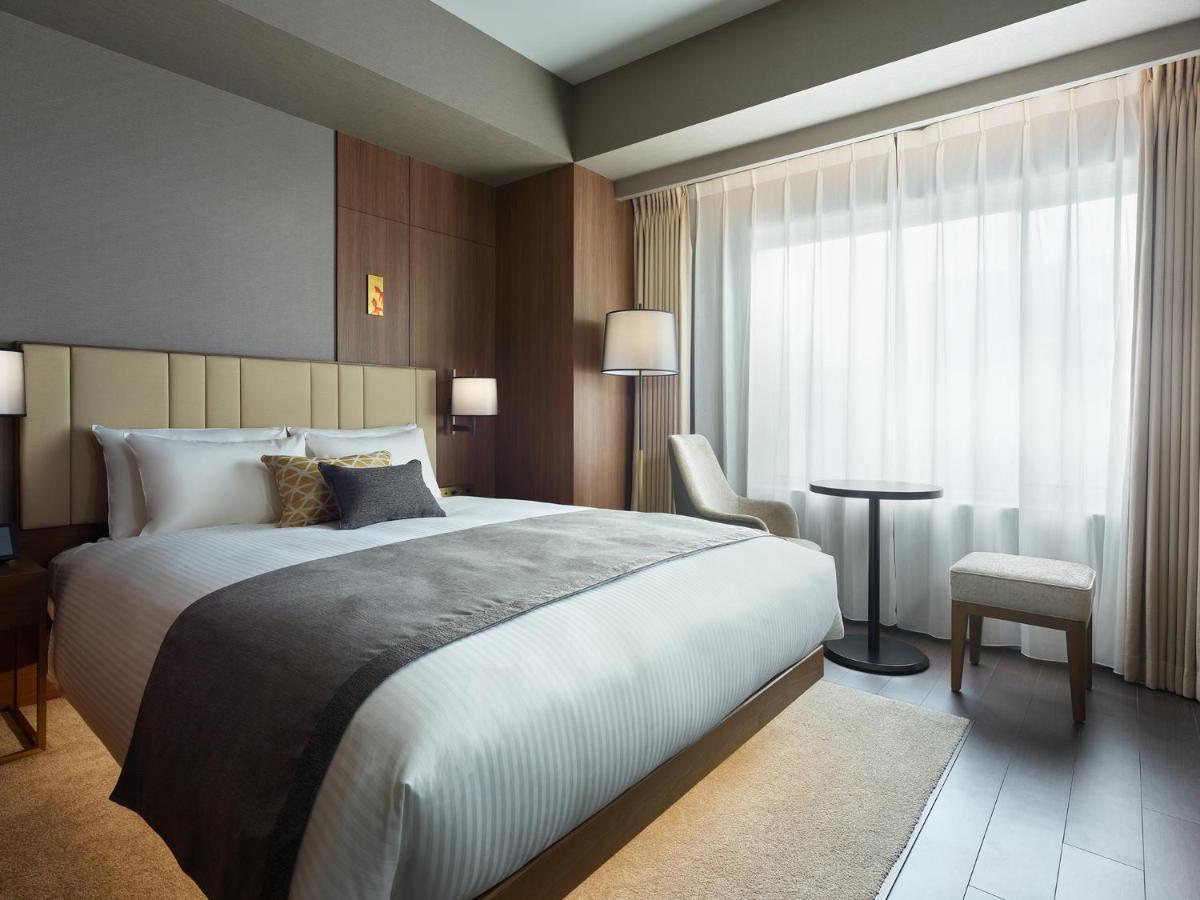 Mitsui Garden Hotel Roppongi Tokyo Premier Екстериор снимка The photo shows a modern hotel room featuring a comfortable bed with white linens and a decorative throw pillow. The room has a neutral color palette with wooden accents on the walls. There are two bedside lamps and a chair positioned next to a small
