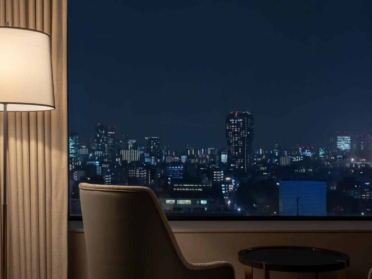 Mitsui Garden Hotel Roppongi Tokyo Premier Екстериор снимка The photo depicts a cozy hotel room setting at night. In the foreground, there's a stylish armchair next to a small round table. A standing lamp provides soft lighting, creating a warm ambiance. In the background, large windows reveal a city skyline 