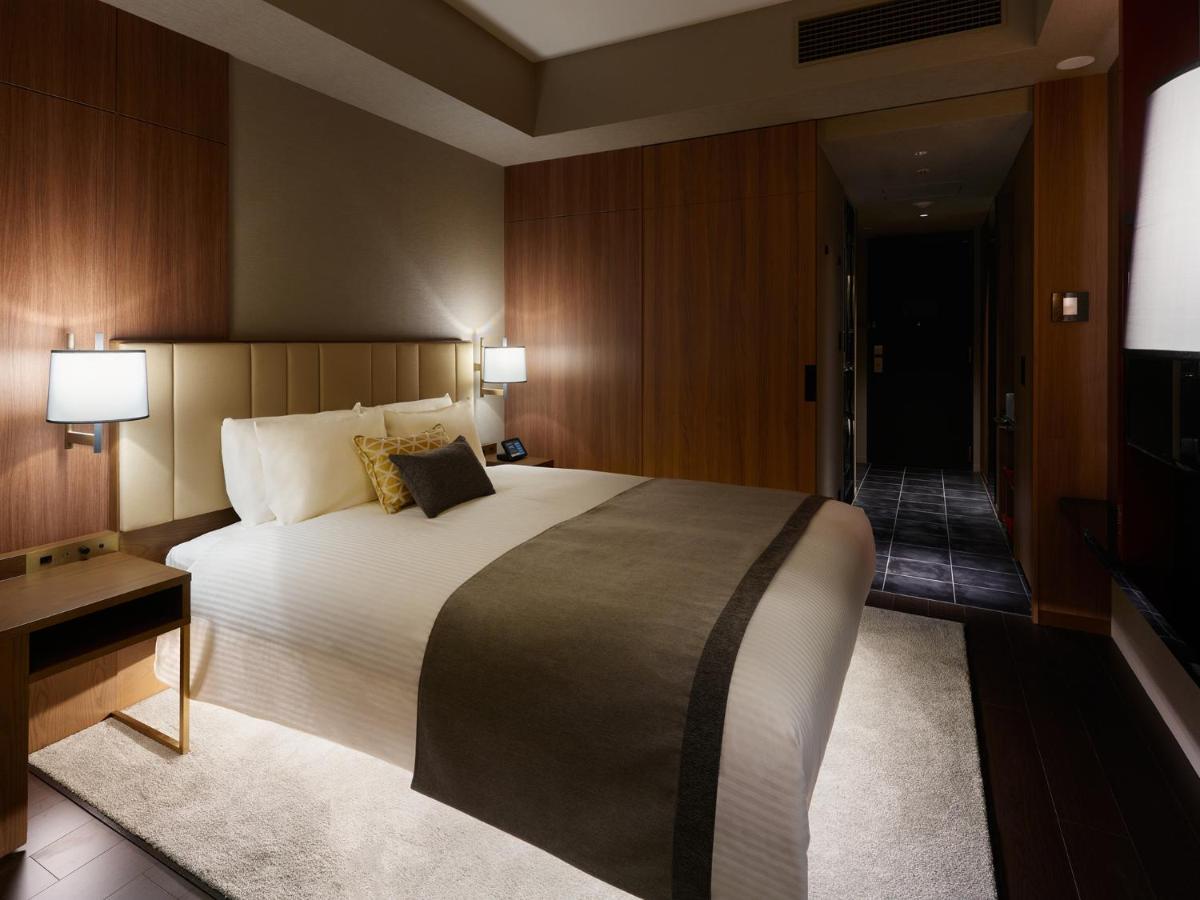 Mitsui Garden Hotel Roppongi Tokyo Premier Екстериор снимка The image shows a modern hotel room with a warm and elegant design. In the center, there is a neatly made bed with white linens and a dark gray throw blanket. Flanking the bed are two bedside lamps providing soft lighting. The walls have a rich wood 