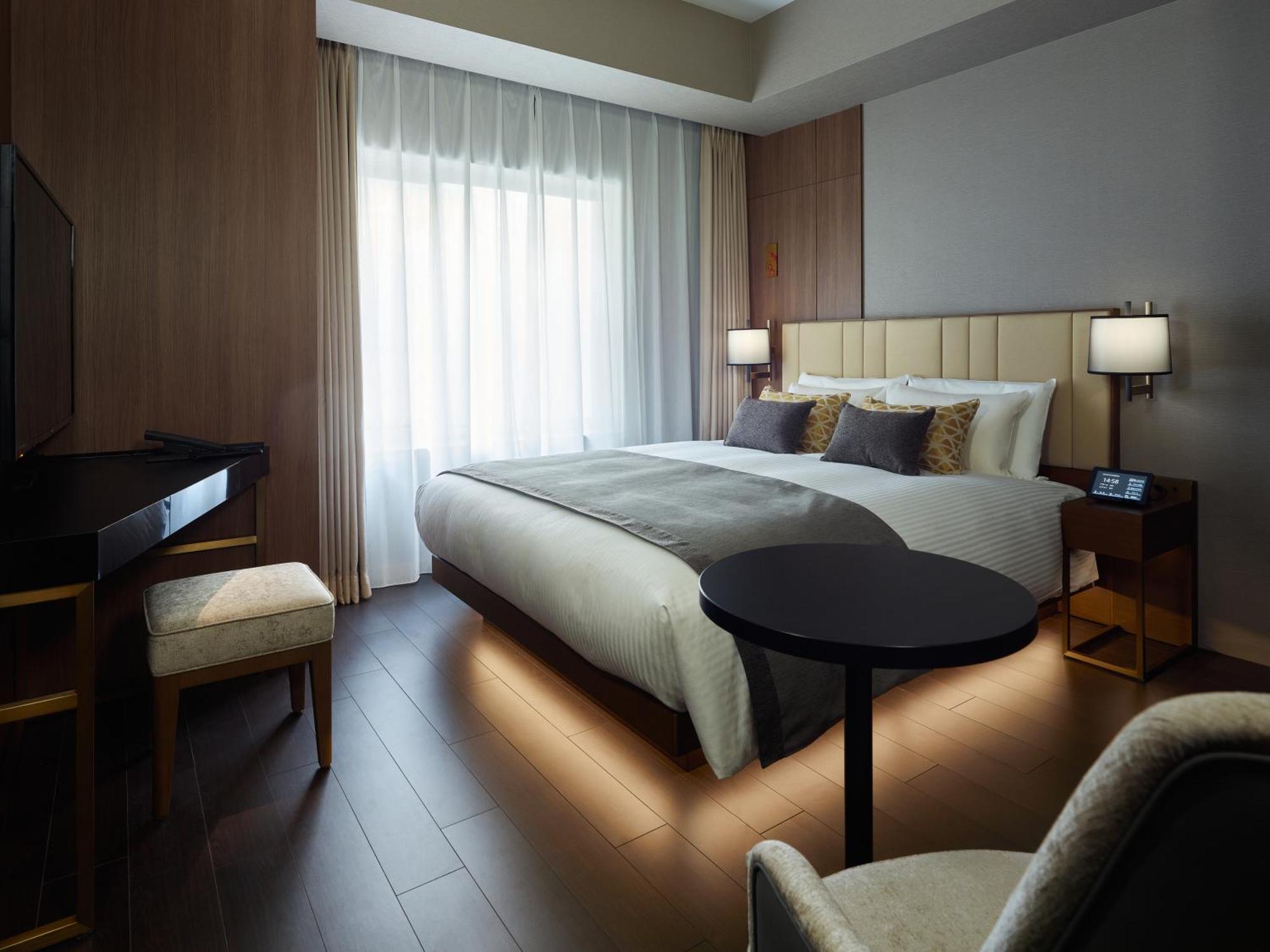 Mitsui Garden Hotel Roppongi Tokyo Premier Екстериор снимка The photo shows a modern hotel room featuring a large bed with decorative pillows. There are large windows with sheer curtains allowing natural light to filter in. To one side, there is a sleek desk with a chair and a small round table in the center 